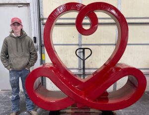 Man standing beside a large red custom fabrication sculpture shaped like a stylized heart and loop