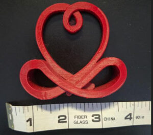 Top-down view of a small red custom fabrication sculpture piece next to a ruler showing 3-inch width